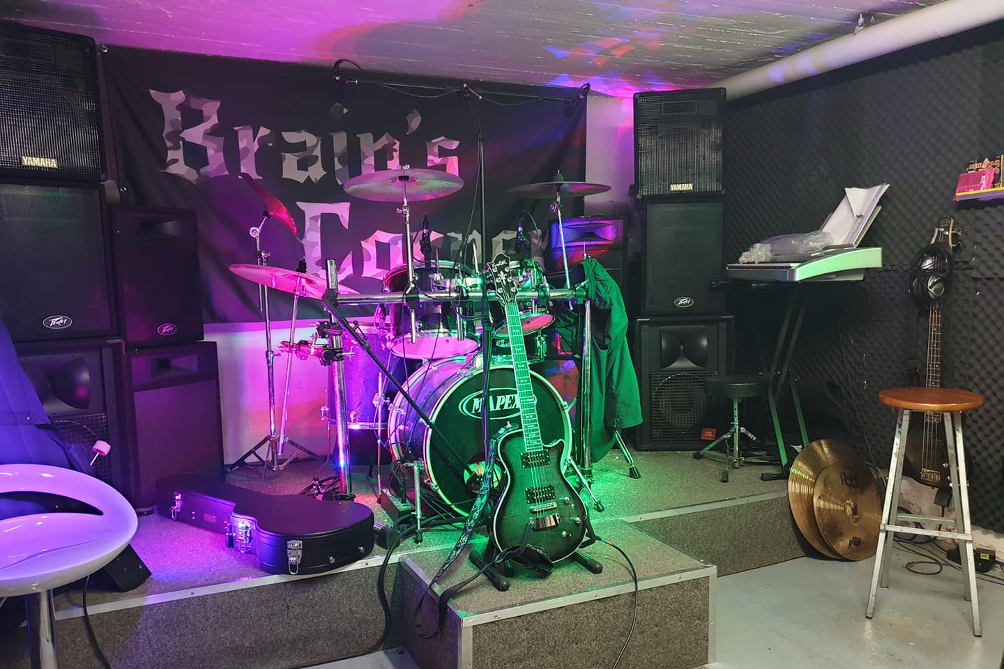 Proberaum von Brain’s Corner mit Schlagzeug, Gitarren und Bandbanner in violettem und grünem Licht